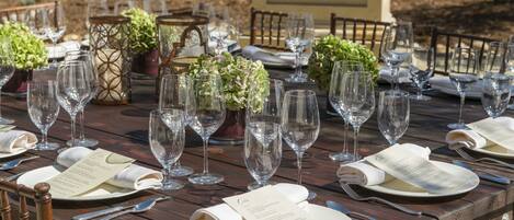 A wooden dining table set with multiple wine glasses, plates, and cutlery, accompanied by menus and decorative greenery.