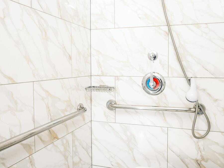 Zona de ducha con baranda, panel de control con un botón rojo y pared de azulejos blancos.