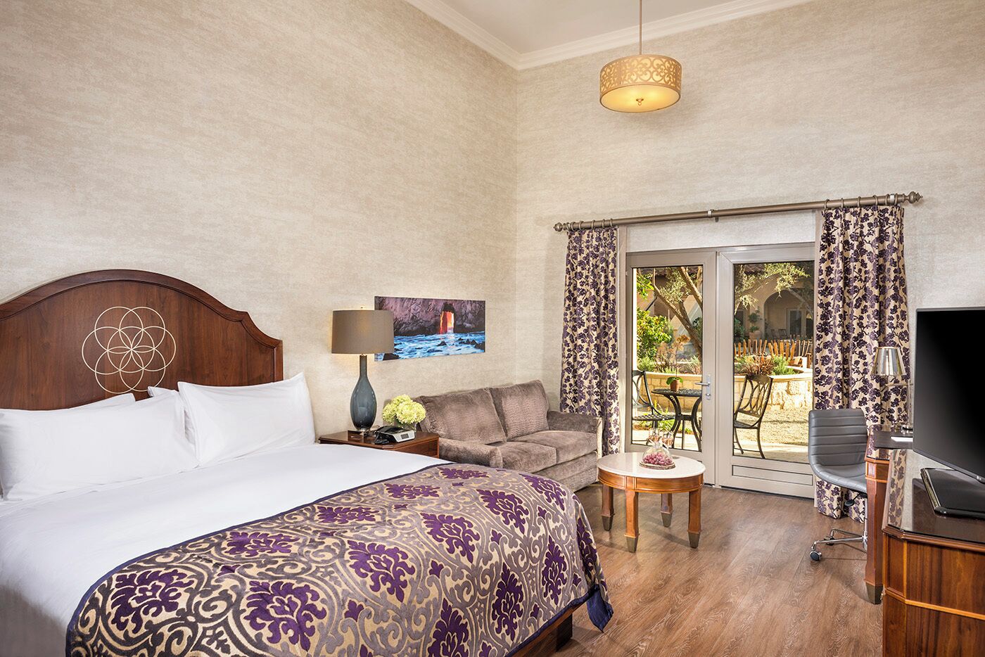 A hotel room with a bed, a sofa, a TV, a table with a vase, and a painting on the wall.