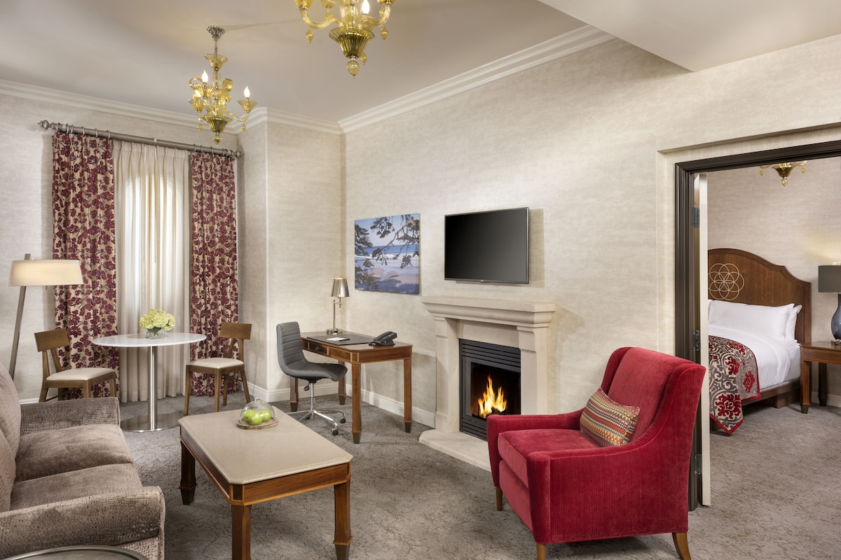 Habitación de hotel con chimenea, televisor en la pared, un sillón rojo, sofá, mesa de comedor con sillas y una mesita con lámpara y flores.