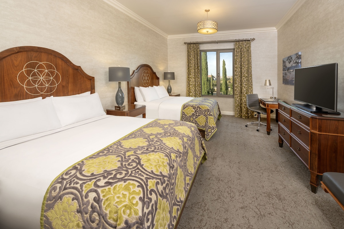 Habitación de hotel con dos camas, televisión, escritorio y ventana con cortinas.