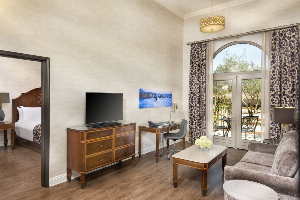 Habitación de hotel con cama, televisión, escritorio y silla, sofá y un ventanal con cortinas.