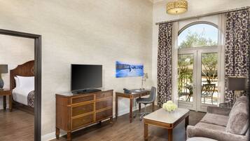Habitación de hotel con cama, televisión, escritorio y silla, sofá y un ventanal con cortinas.