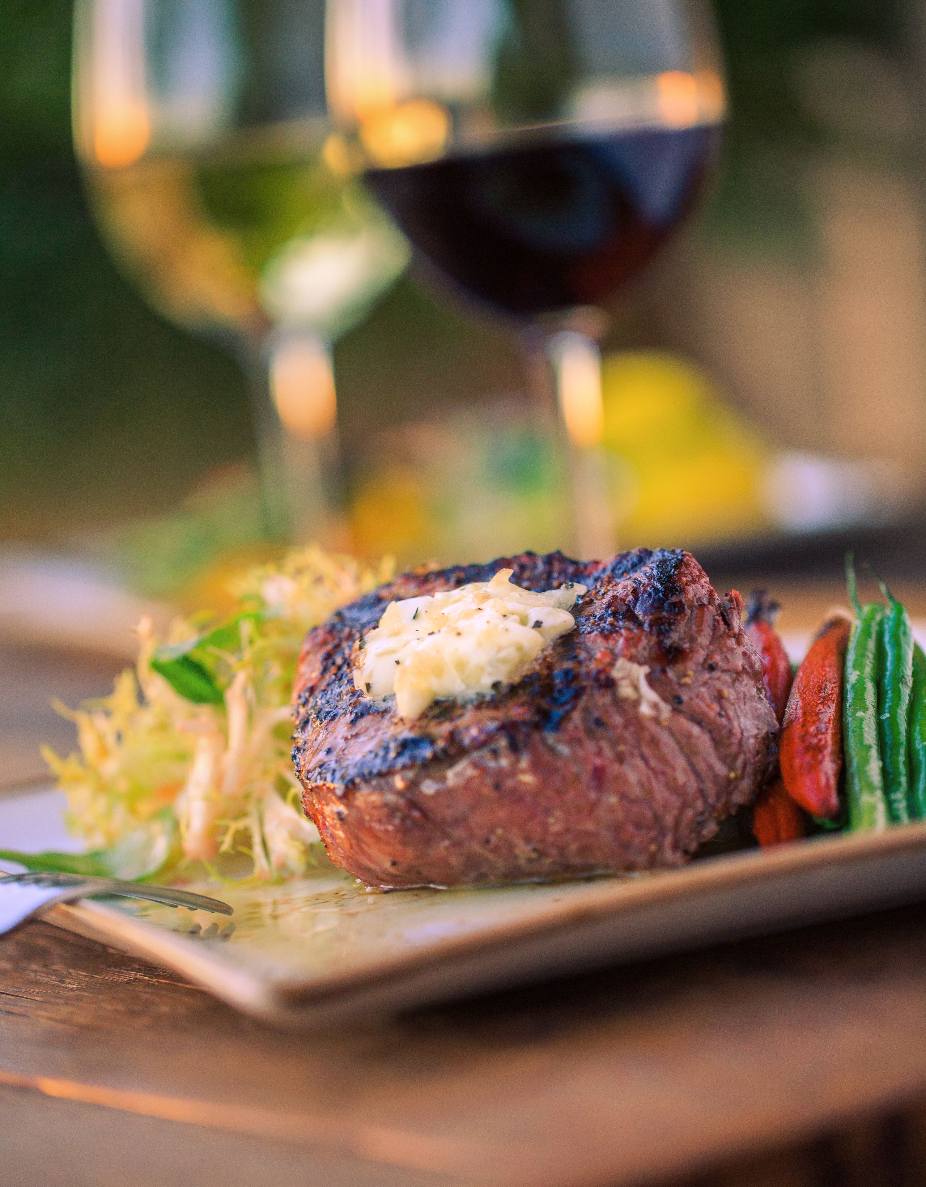 A steak with a pat of butter on top, served with vegetables and a glass of red wine in the background.