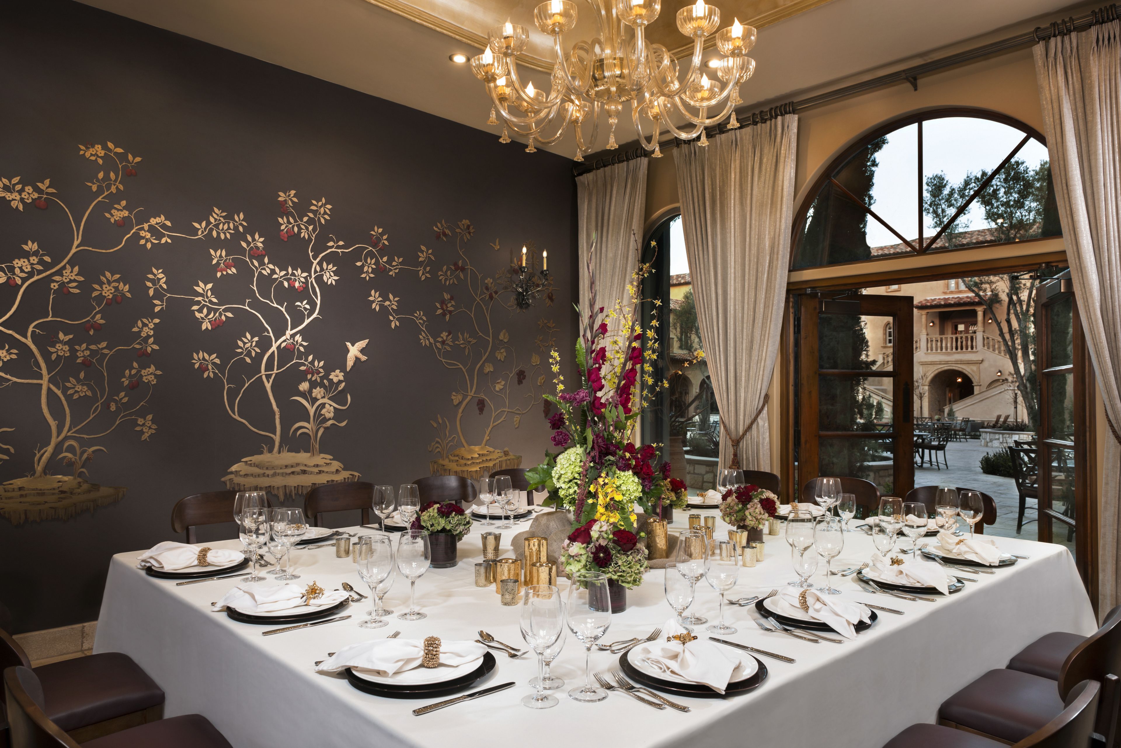 Comedor con una larga mesa dispuesta para una comida, con un candelabro, arreglos florales y vistas al exterior.