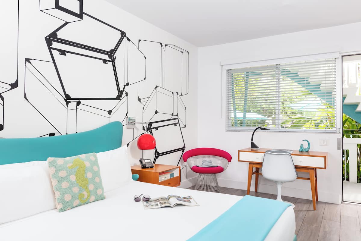 A modern bedroom with a bed, desk, and chair, a geometric wall design, and a window with blinds.