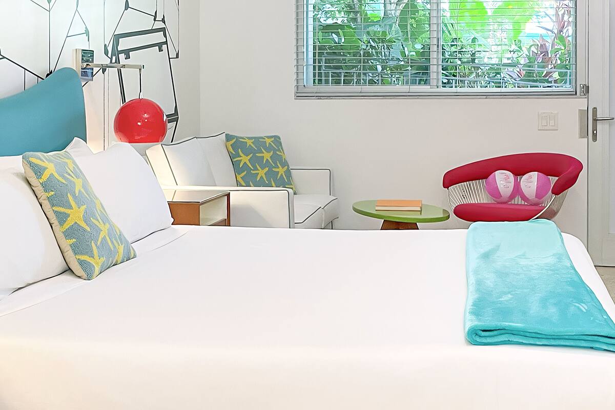 A modern bedroom with a bed, a small table, a chair, and a window with a view of greenery.