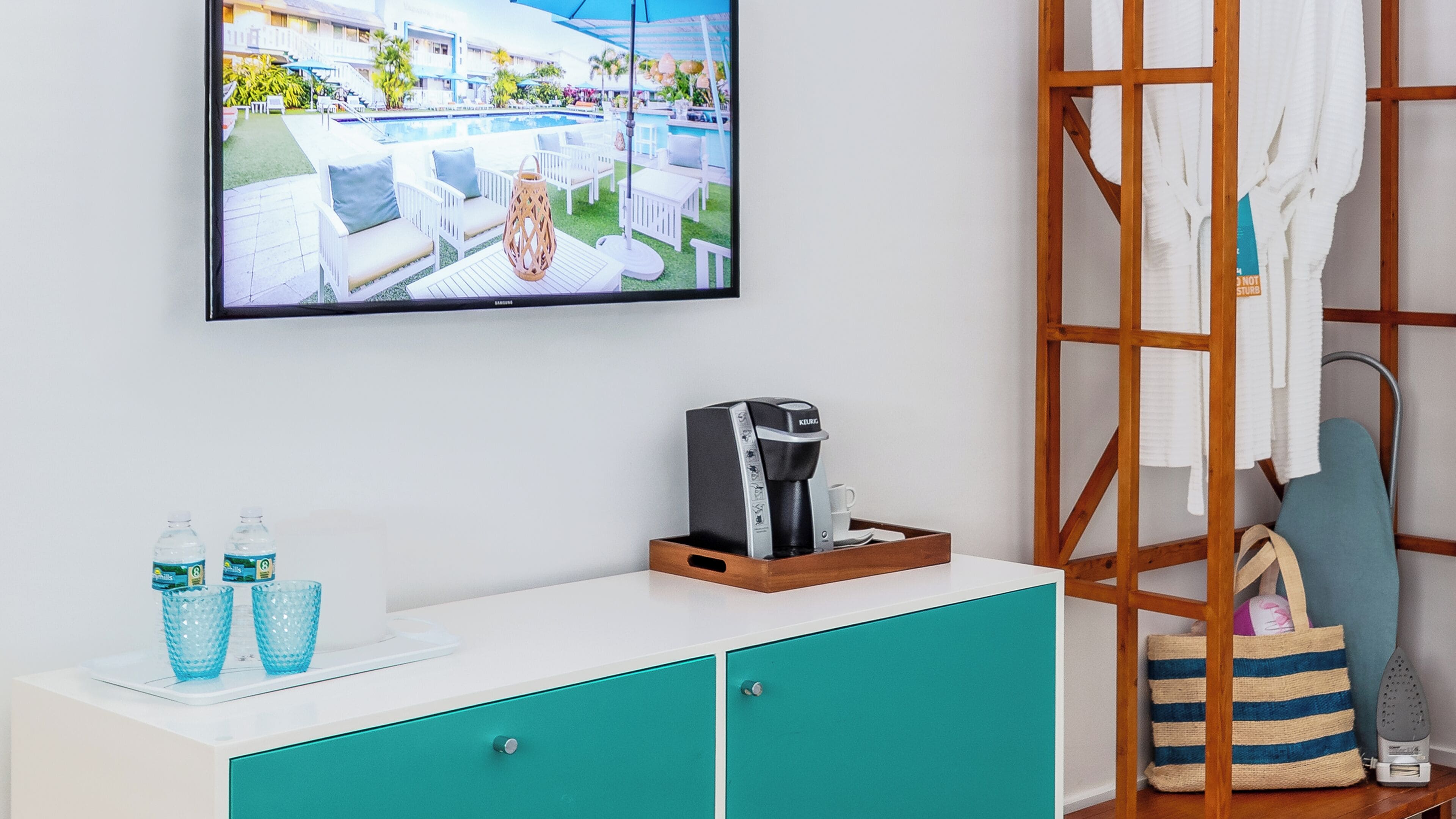 A hotel room with a television displaying a pool view, a coffee maker, a bottle of water, and a striped bag.
