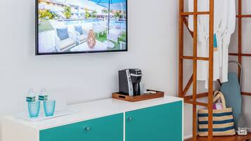 A hotel room with a television displaying a pool view, a coffee maker, a bottle of water, and a striped bag.