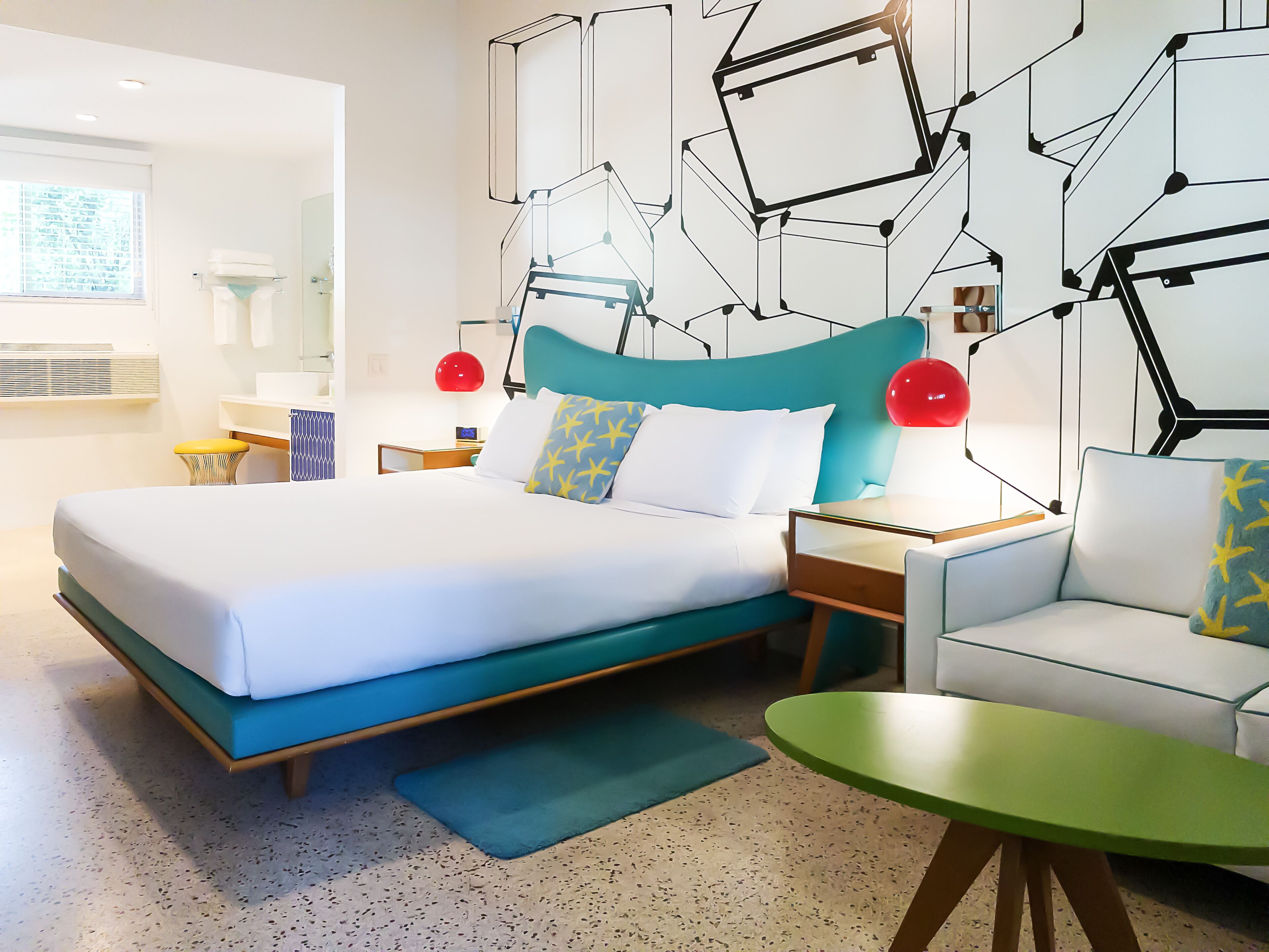 A modern hotel room with a large bed, a green armchair, a small table, and a geometric wall design.