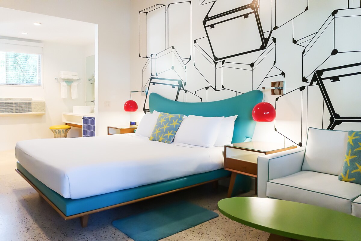 A modern hotel room with a large bed, a green armchair, a small table, and a geometric wall design.
