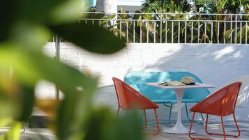 Een patio met een witte bakstenen muur, een blauwe bank, twee oranje stoelen en een tafeltje met een hoed en krant.