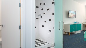 A hotel room with a checkered floor, a shower with white and black tiles, a teal cabinet, and a television on the wall. - The Vagabond Hotel Miami (Miami)