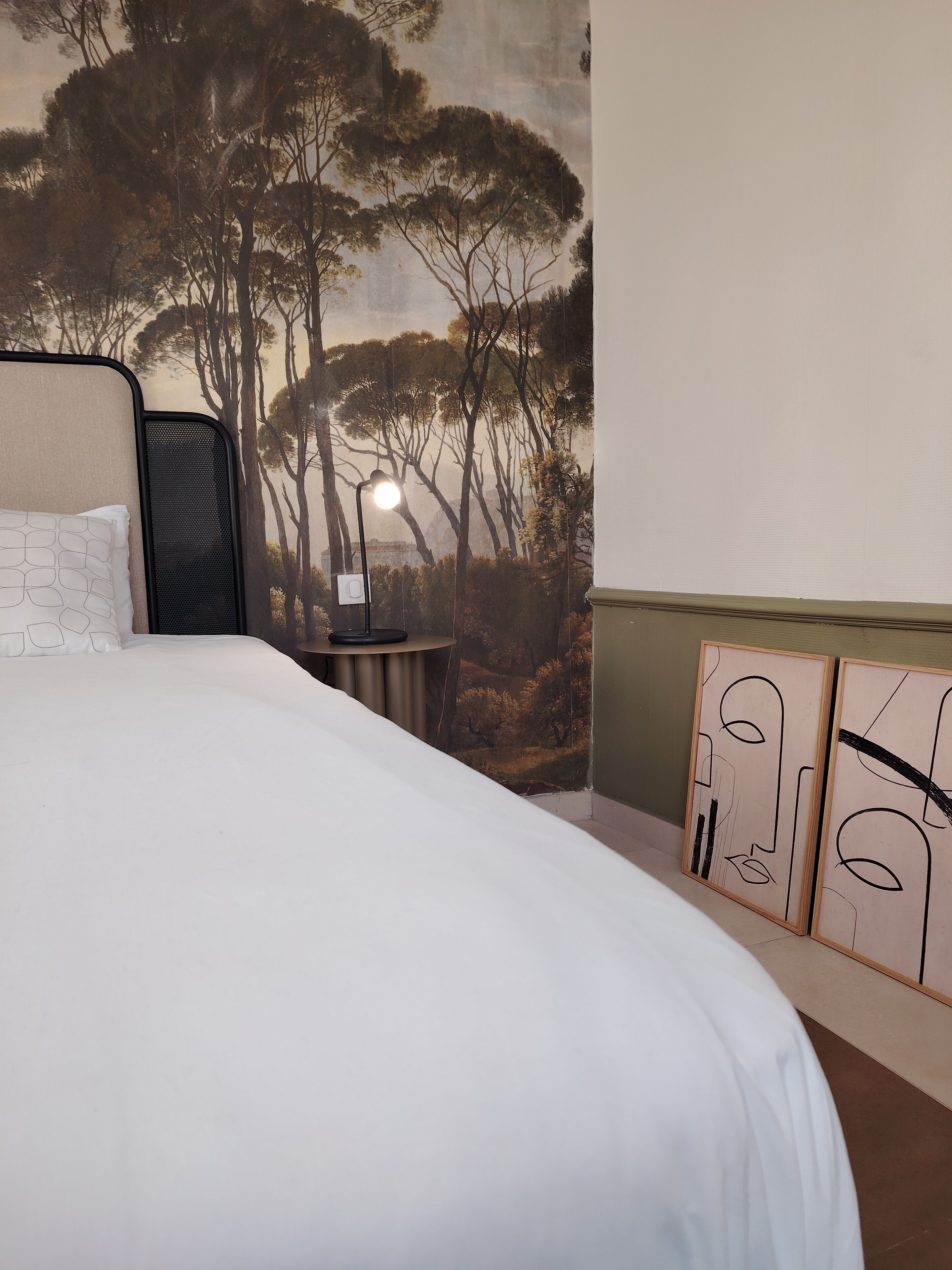 A bedroom with a bed, a nightstand, a lamp, and a wall mural of trees.