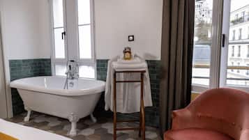 Une salle de bain avec une baignoire sur pieds, un tabouret en bois et une vue sur un immeuble par la fenêtre.