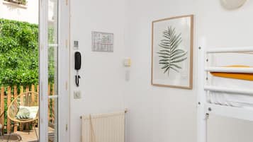 A room with a wooden ceiling, a white radiator, a wicker chair, a small stool, a framed plant, and a bunk bed.