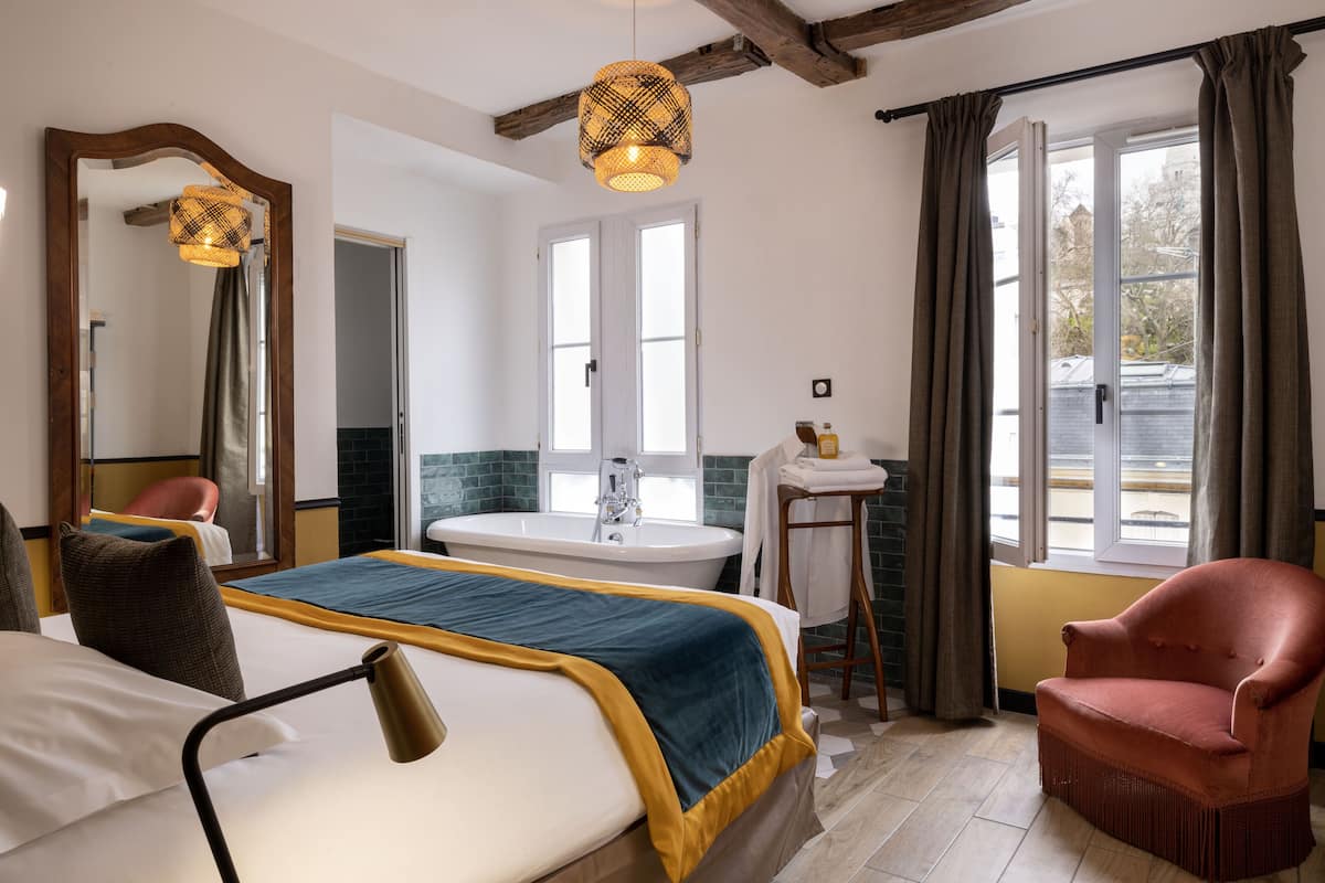 Een slaapkamer met een bed, een stoel, een badkamer met een wastafel en spiegel, en een raam met gordijnen.