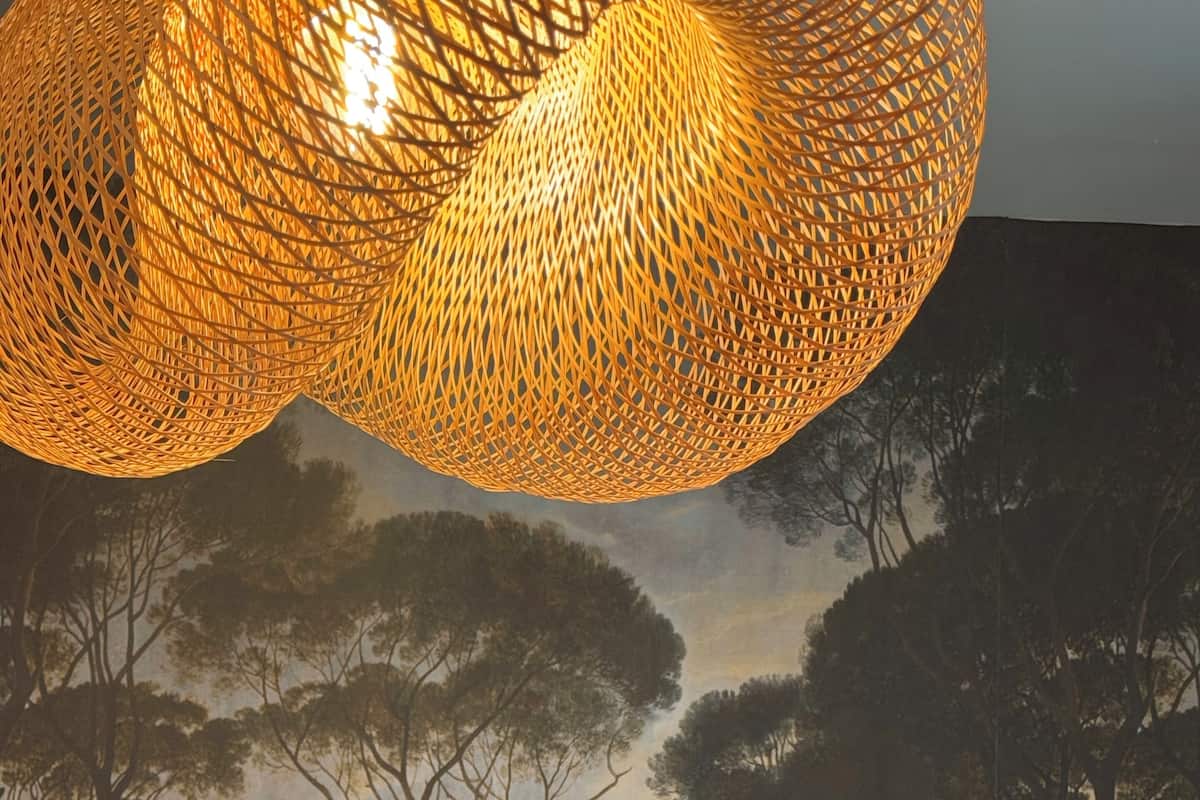 Een slaapkamer met een bed, gestreepte kussens, een textuurrijk behang en een geweven lamp.