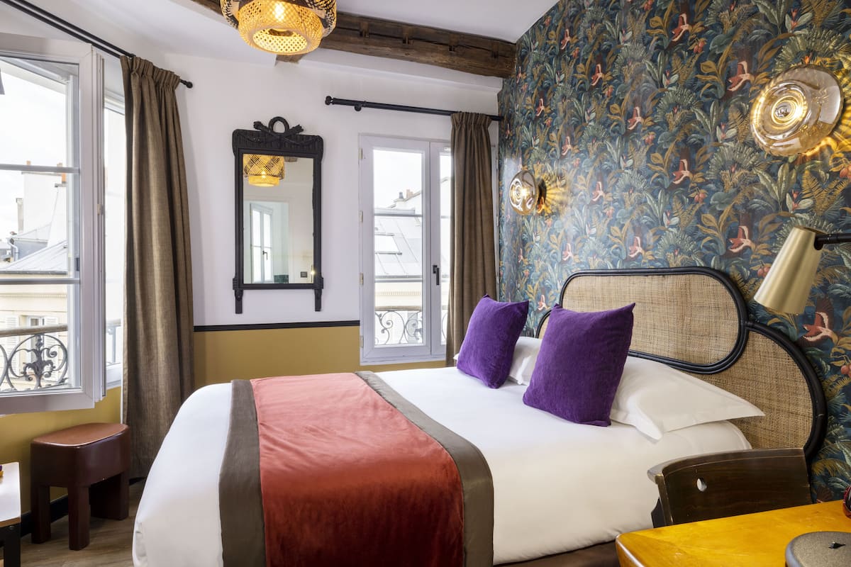 Hotelkamer met een groot bed, een houten hoofdbord, een raam met gordijnen, een klein krukje en een tafel met een lamp.