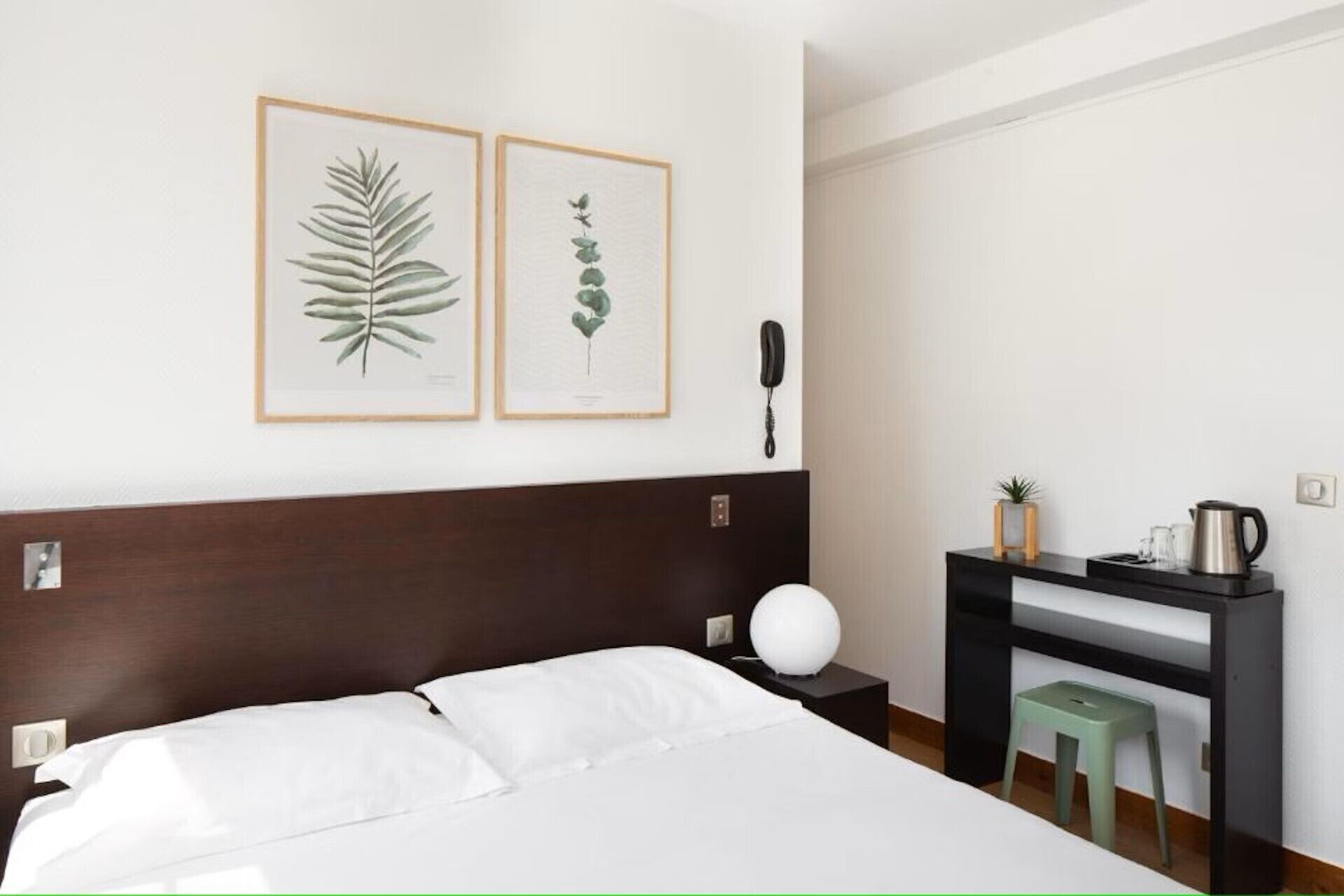 A hotel room with a bed, a nightstand, a desk, a phone, and two framed botanical prints on the wall.