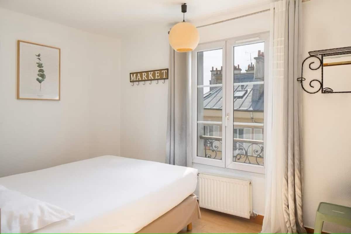 Een slaapkamer met een bed, een raam met uitzicht op gebouwen, een radiator en een wandlamp.