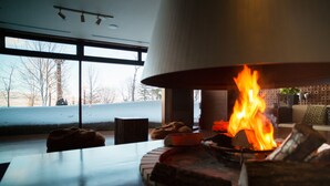 A modern living room with a large fireplace, a wooden table, and a view of a snowy landscape through large windows. - Lake Akan Tsuruga Wings (Kushiro)