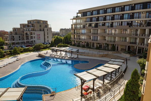 A swimming pool with a wavy design, surrounded by lounge chairs and umbrellas, with apartment buildings in the background. - Menada Luxor Apartments (Sveti Vlas)