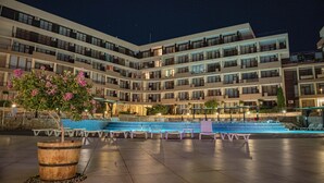 Un hotel de varios pisos con balcones, un área de piscina con sillas de descanso y una planta grande en maceta con flores rosas.