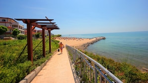 Un paseo marítimo con barandilla metálica que conduce a una zona de playa con gente y un muelle.