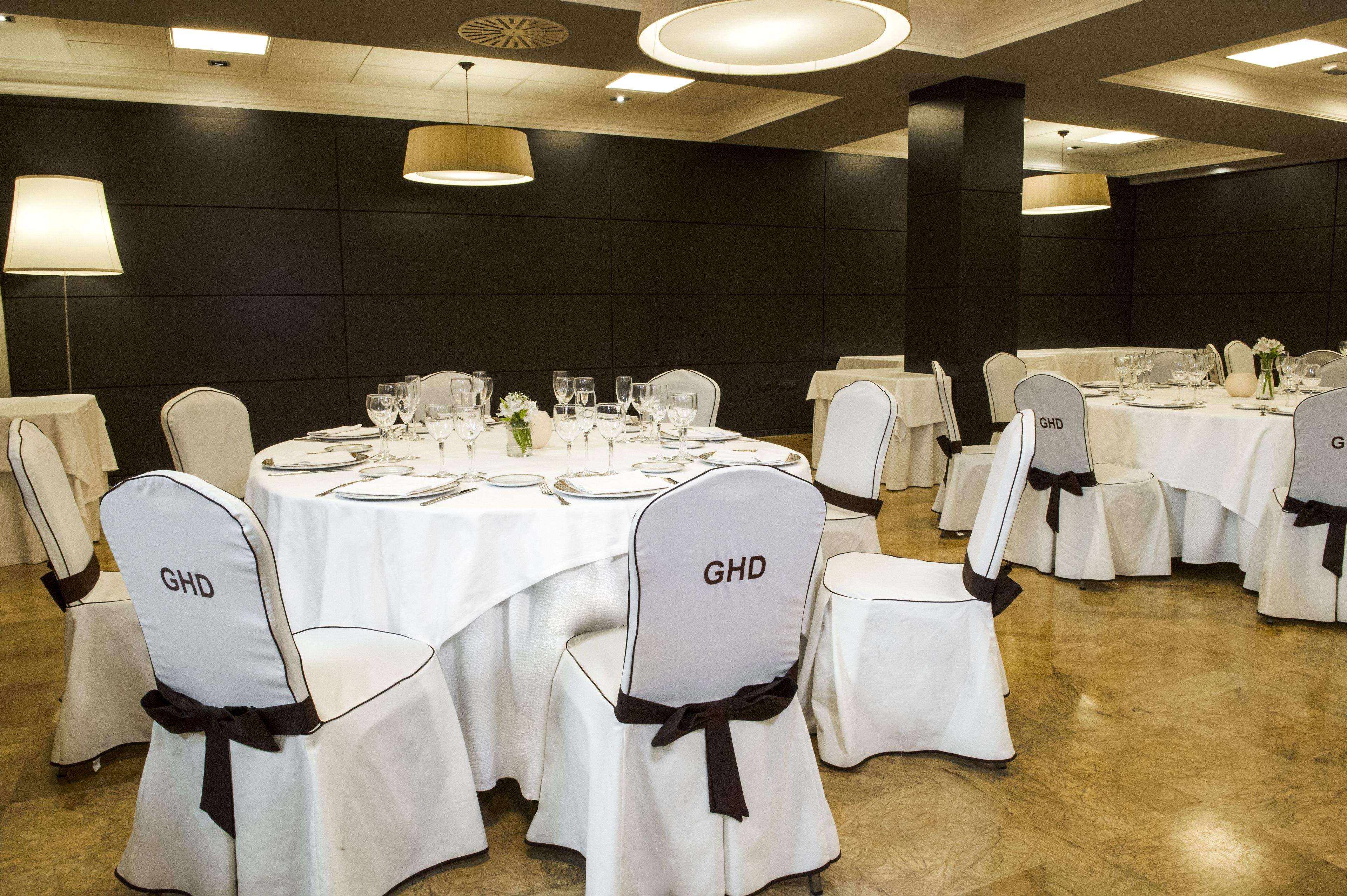 Un salón de banquetes con mesas redondas dispuestas para comer, cada una con manteles blancos y sillas con el logo 'GHD'.