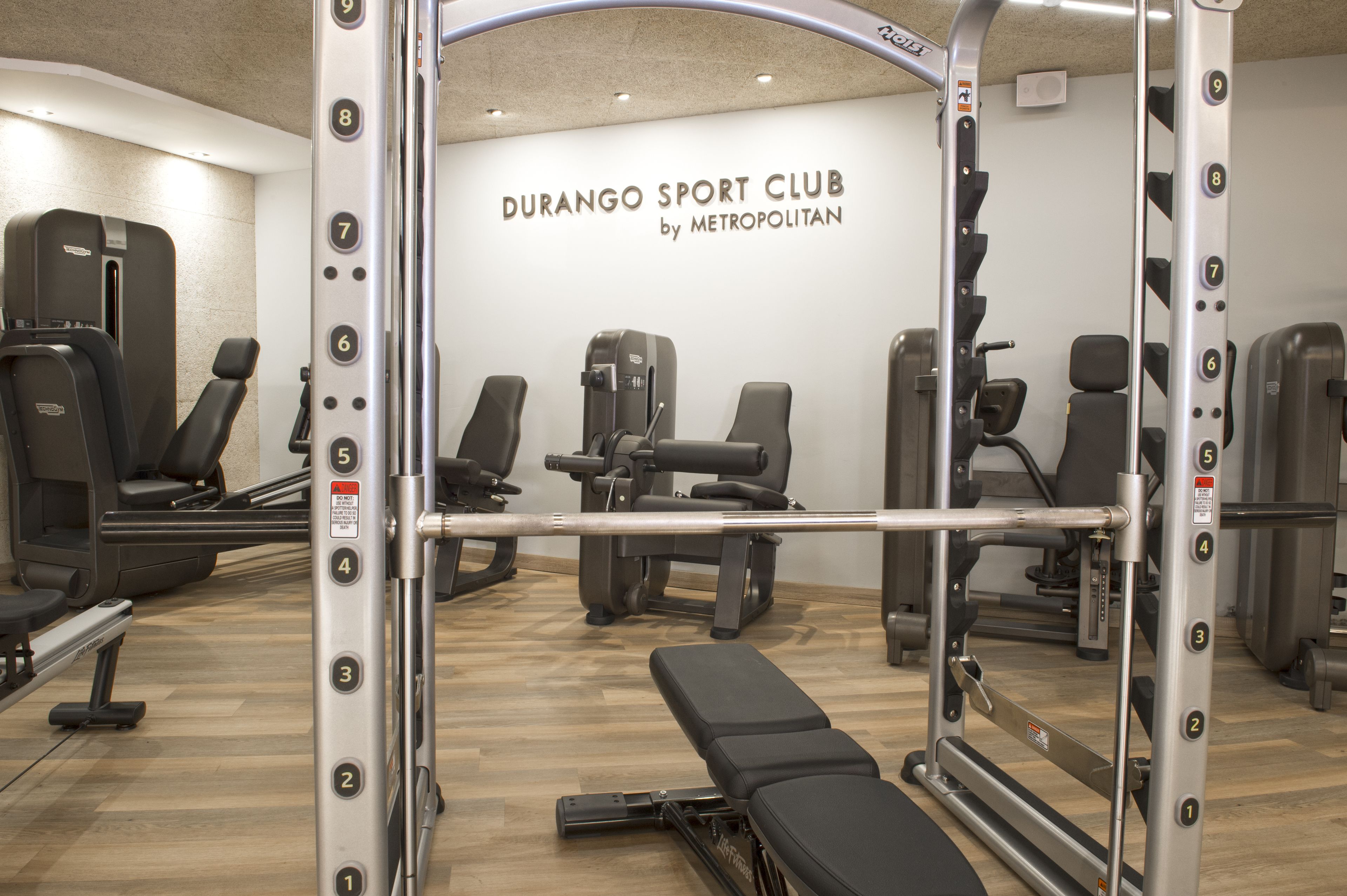 Un gimnasio con diversos equipos de ejercicio, incluyendo máquinas de pesas y bancos, dispuesto en un espacio amplio.