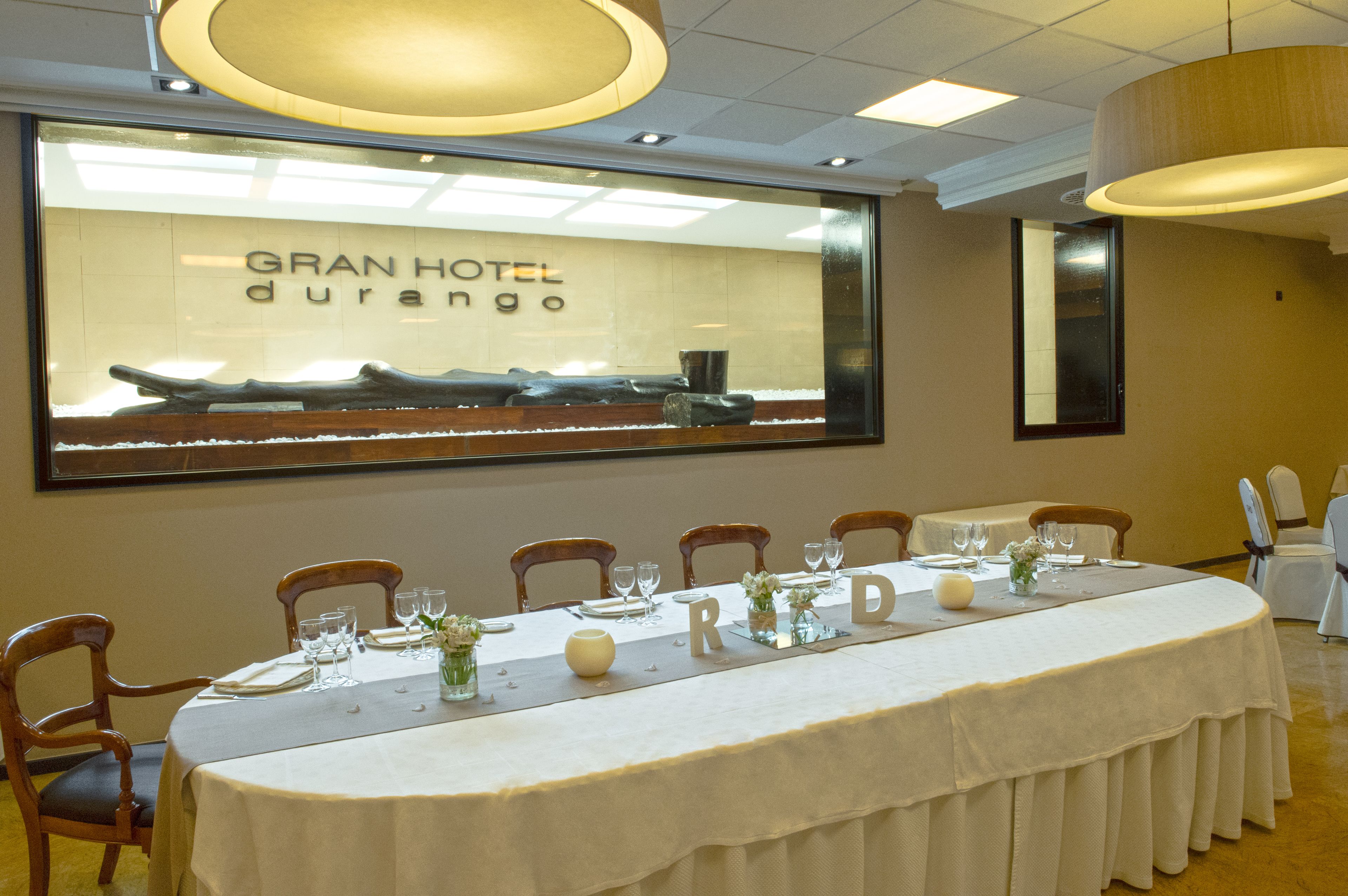 A hotel room with a long dining table set for a meal, a large window with the hotel's name, and a modern chandelier.