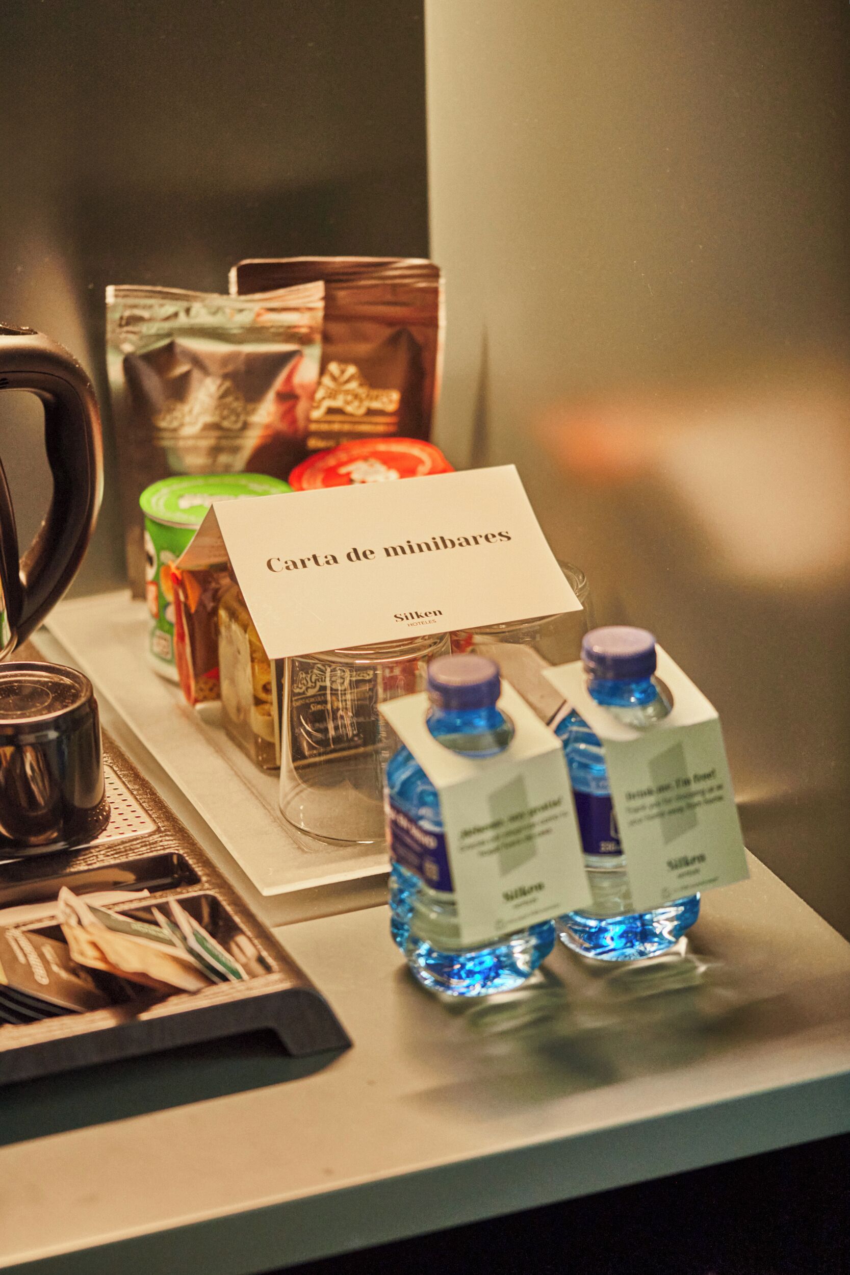 Un minibar con agua embotellada, una cafetera y diferentes alimentos.