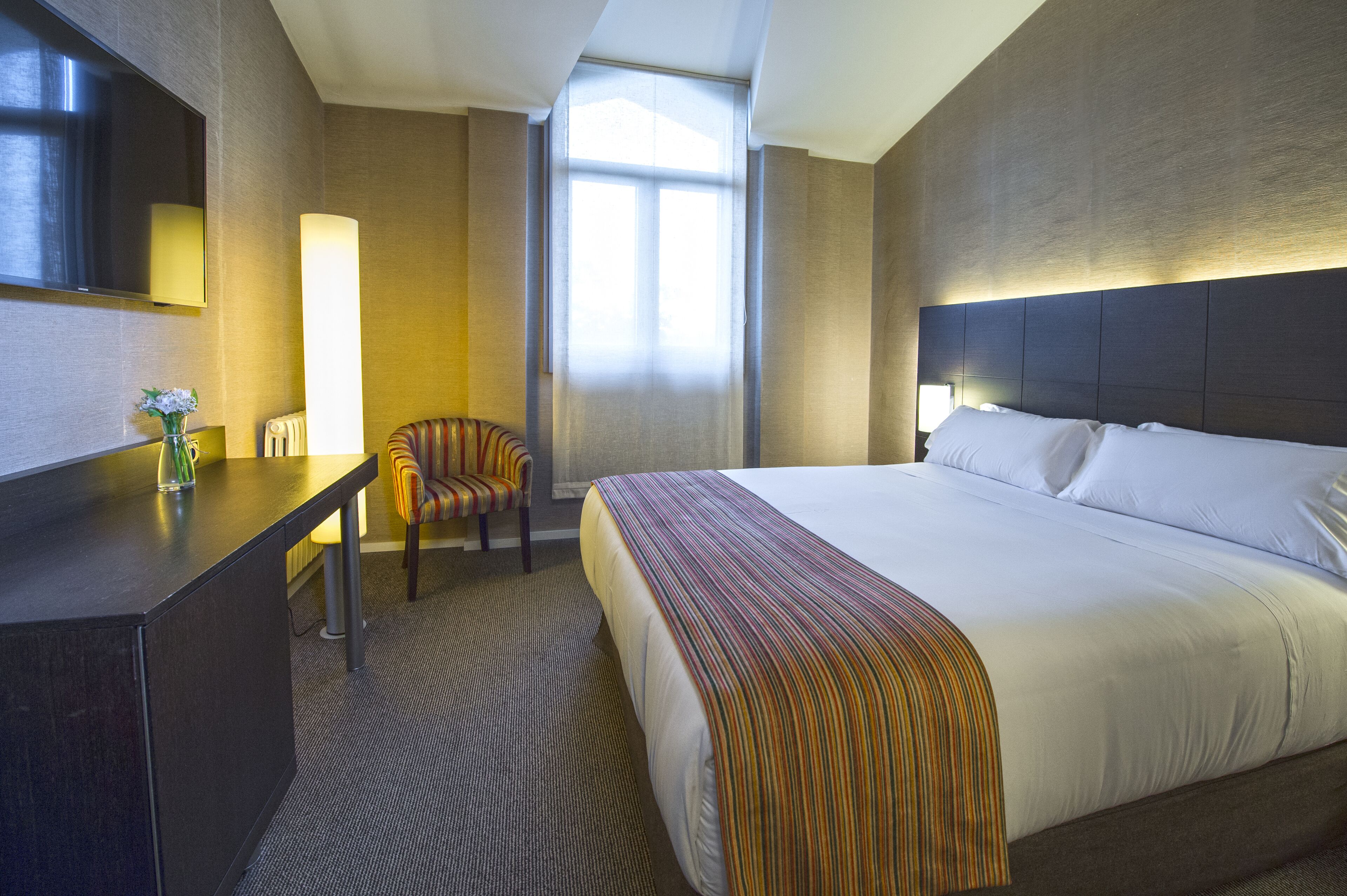 Habitación de hotel con una cama grande, un escritorio con lámpara, una silla y una ventana con cortinas.