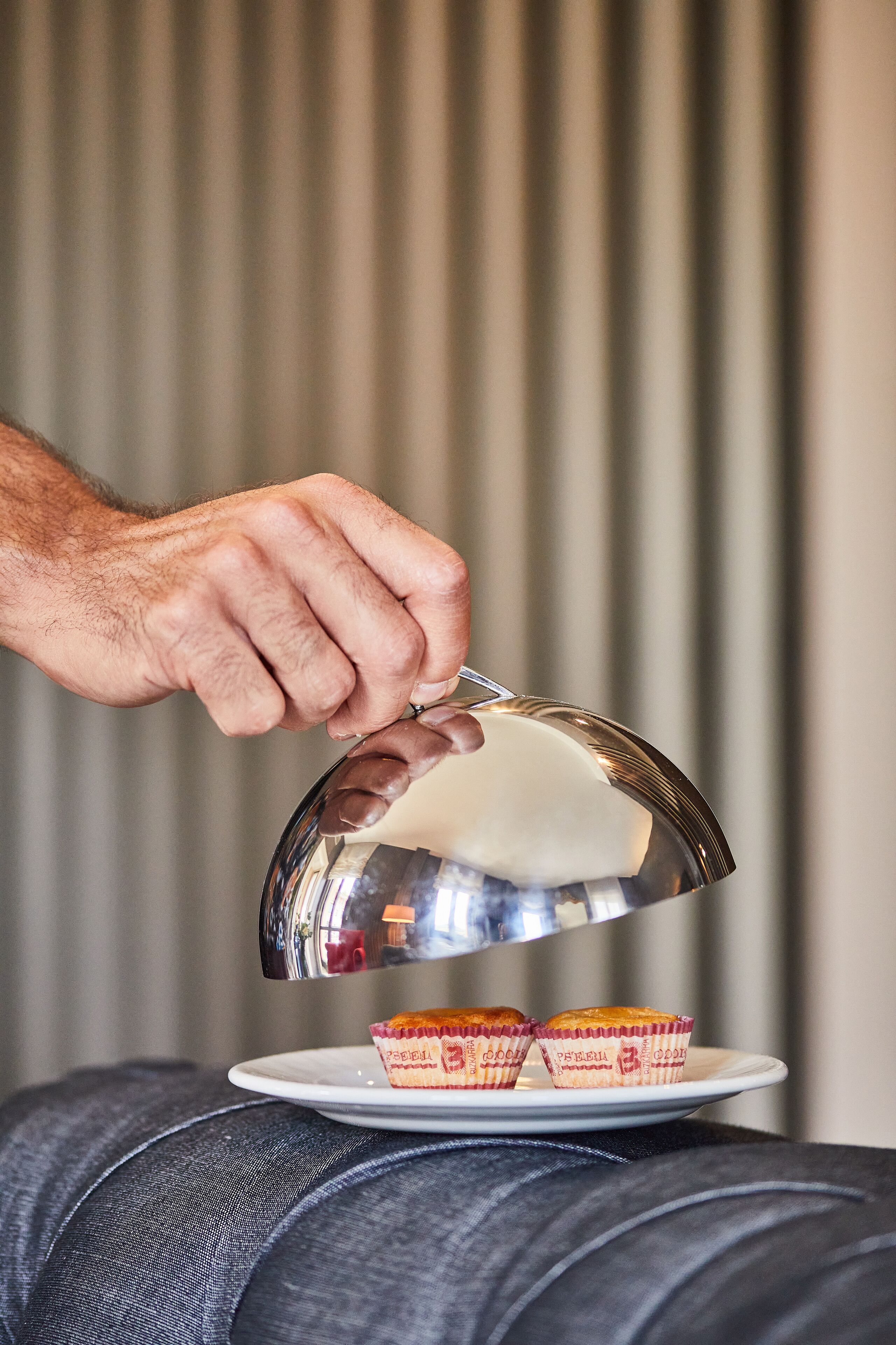 A hand holding a silver teapot over a plate with two cupcakes.