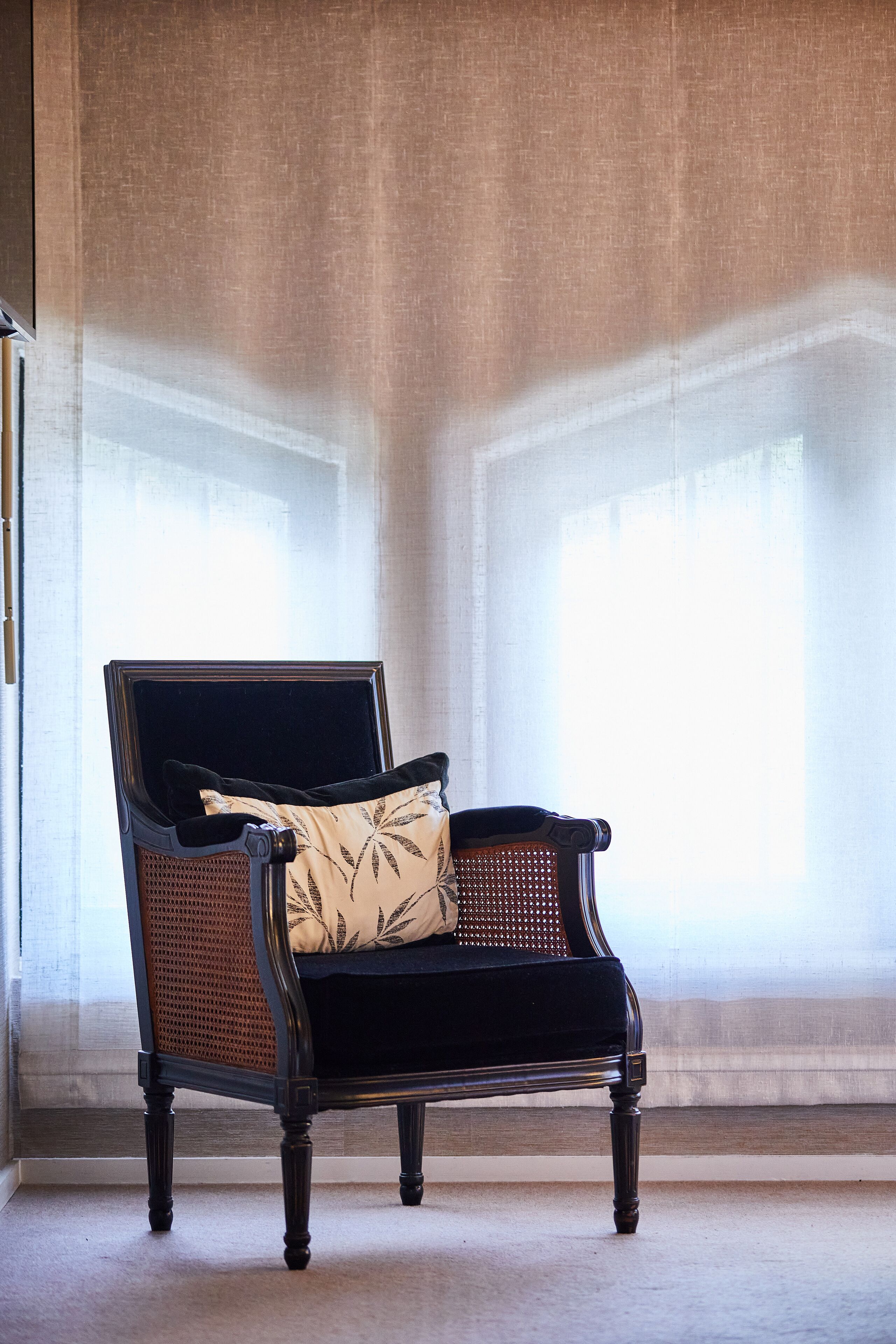 A black upholstered armchair with a woven backrest and a cushion, positioned in front of a window with sheer curtains.
