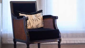 A black upholstered armchair with a woven backrest and a cushion, positioned in front of a window with sheer curtains.