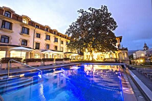 Une piscine à l’eau d’un bleu limpide, entourée d’un bâtiment doté de nombreux balcons et fenêtres, et d’un grand arbre au centre.
