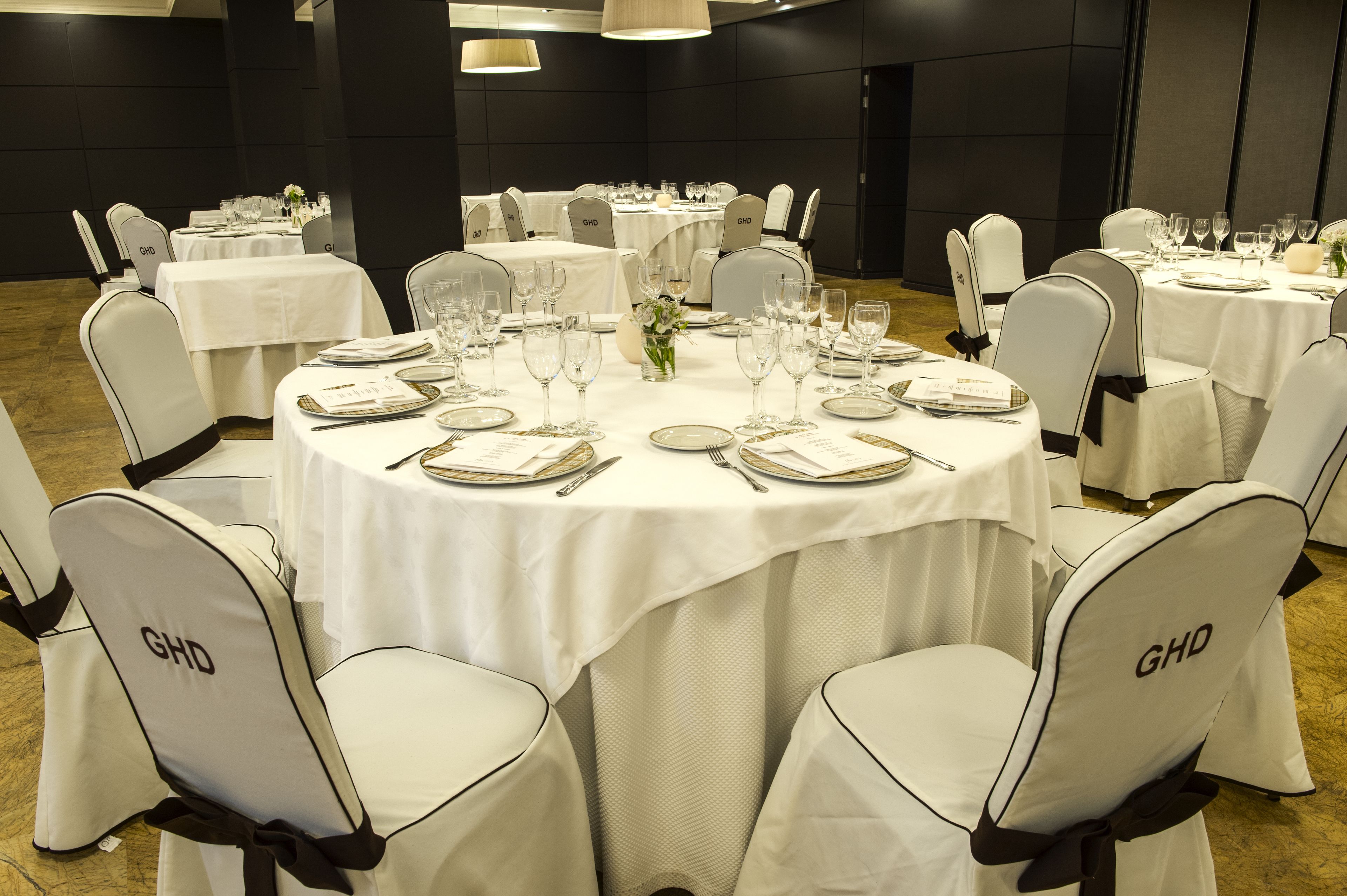 Une salle de banquet avec des tables rondes dressées pour le repas, chacune recouverte d’une nappe blanche et dotée de chaises portant l’inscription « GHD ».