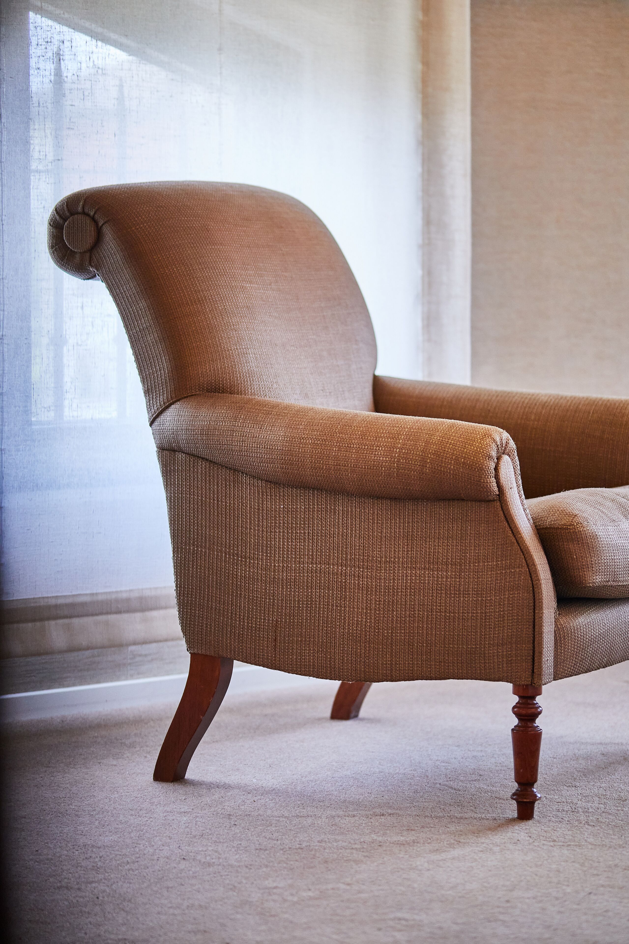 Un sillón marrón con patas de madera en una habitación con una pared lisa y una ventana.