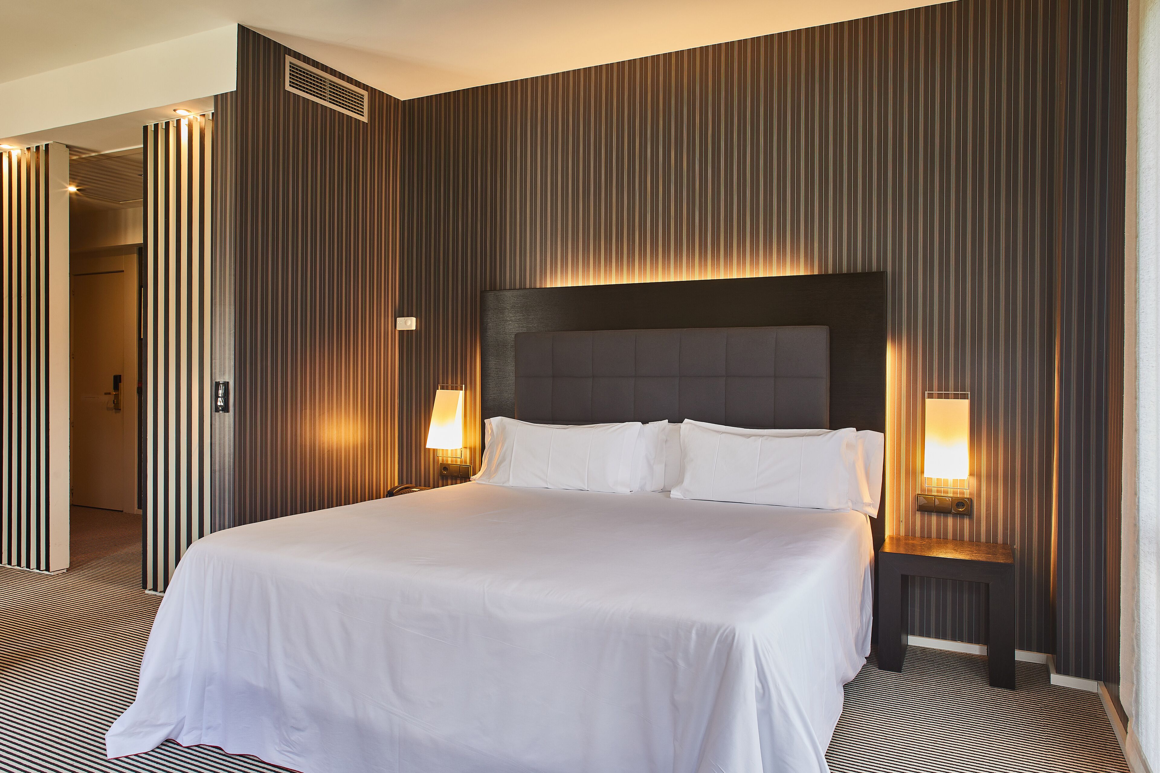 A hotel room with a large bed, two bedside lamps, and a striped wall design.