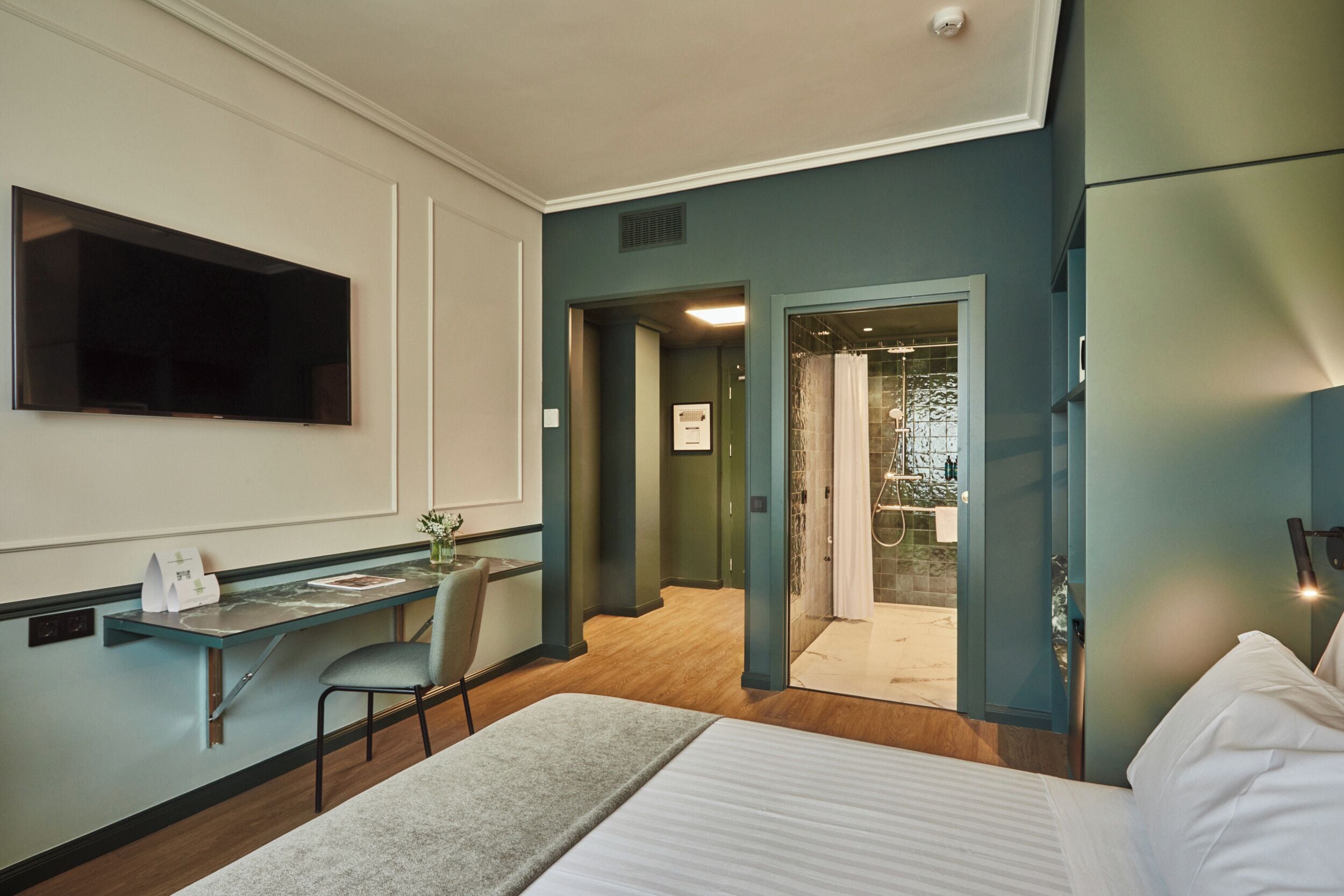 A hotel room with a bed, a desk, a chair, a TV, and a bathroom visible through an open door.