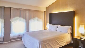A hotel room with a large bed, a bedside table with a lamp, and a view through a window with sheer curtains.