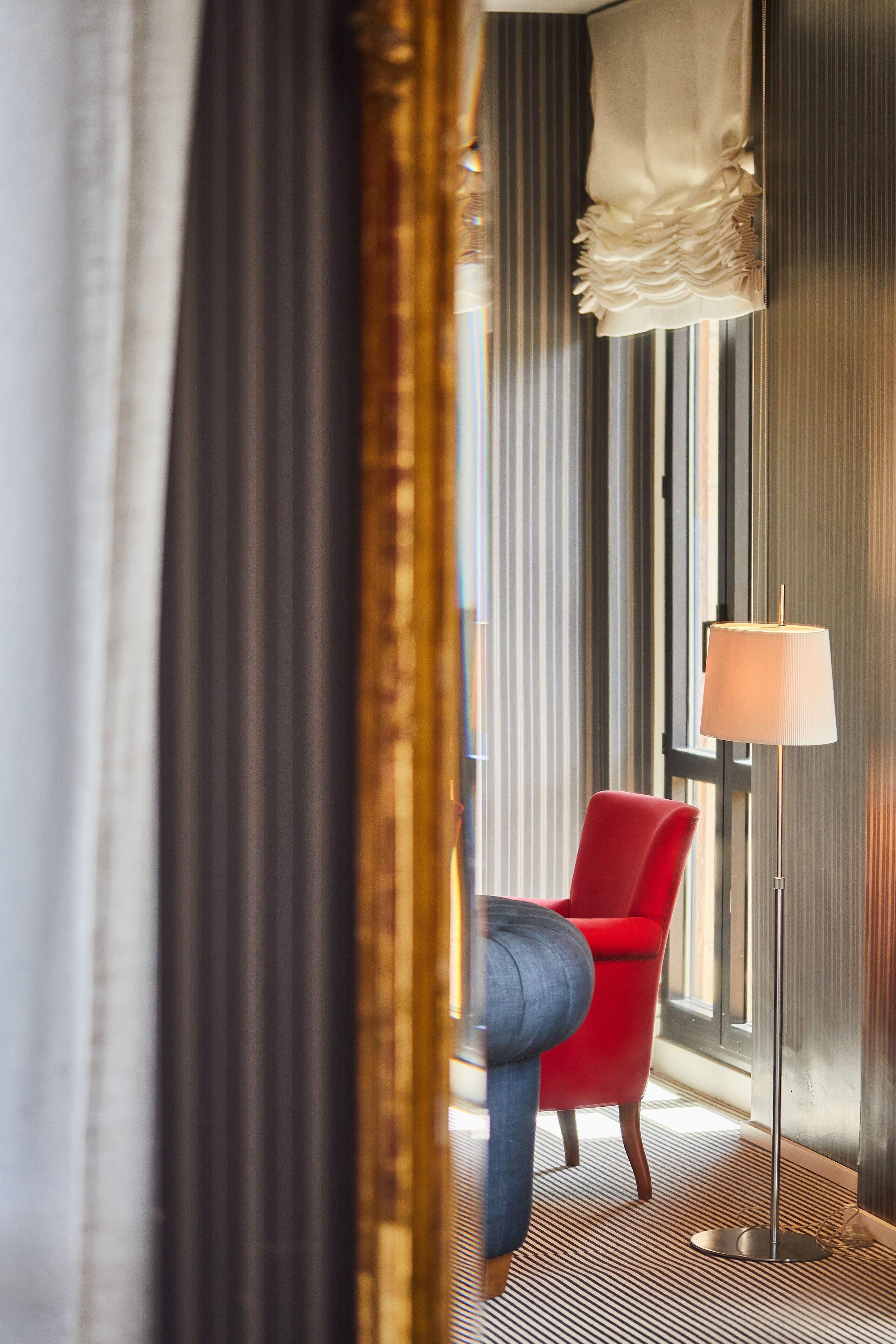A hotel room with a red chair, a floor lamp, and a large window with curtains.
