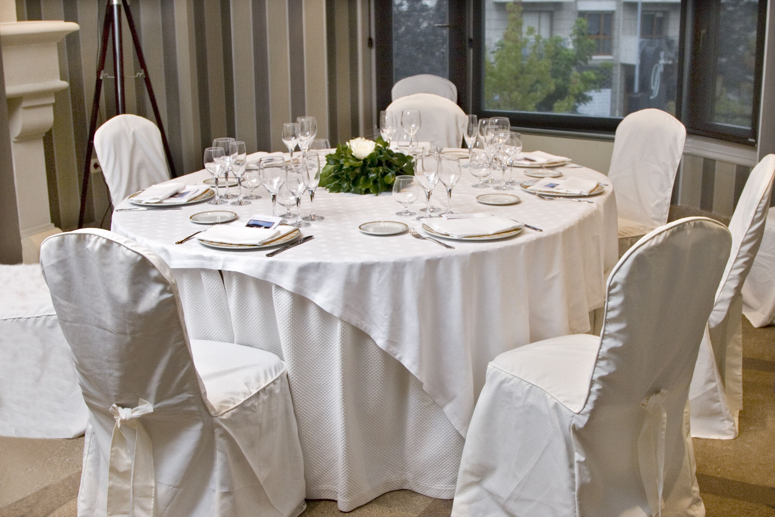 Una mesa redonda dispuesta para un evento formal, con manteles blancos, cristalería y platos.