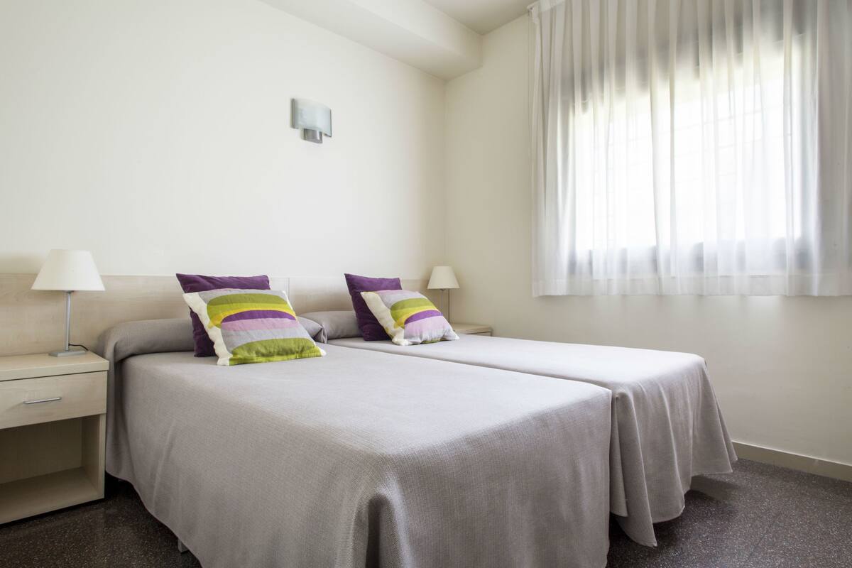 A hotel room with two beds, a nightstand, a lamp, and a window with curtains.