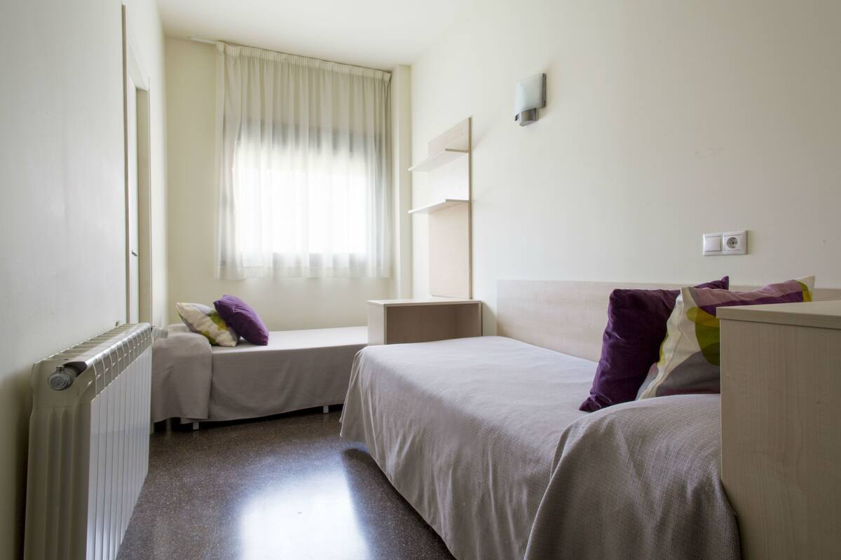 A hotel room with two single beds, a window with curtains, a radiator, and a wall-mounted shelf.