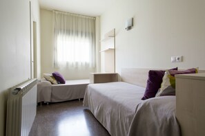 A hotel room with two single beds, a window with curtains, a radiator, and a wall-mounted shelf.