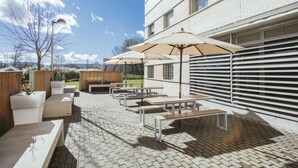 An outdoor patio with white brick flooring, multiple tables and benches, and large umbrellas providing shade. - Albergue Residencia Universitaria Resa Los Abedules - Campus Accomodation (Pamplona)