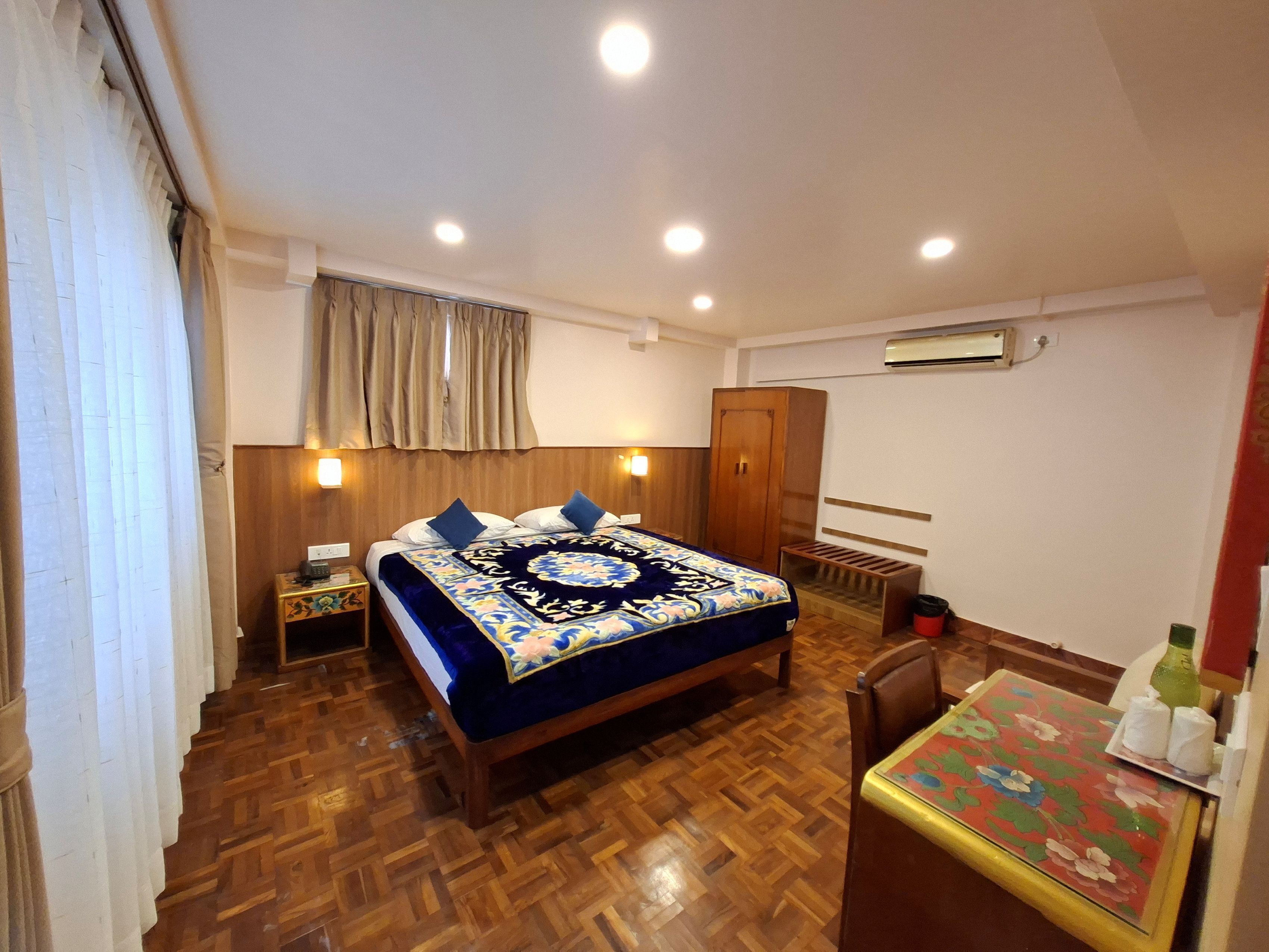 A hotel room with a wooden floor, a bed with a floral bedspread, a wardrobe, a table with a floral tablecloth, and an air conditioning unit.